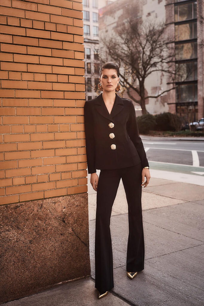 A woman with slicked-back hair leaning against a brick corner on a city sidewalk, showcasing a powerful old money aesthetic from The Boardroom Beauty.jpg. She is wearing a tailored black single-breasted blazer with large, ornate gold statement buttons, matching black wide-leg trousers, pointed-toe gold metallic heels, and chunky gold earrings.