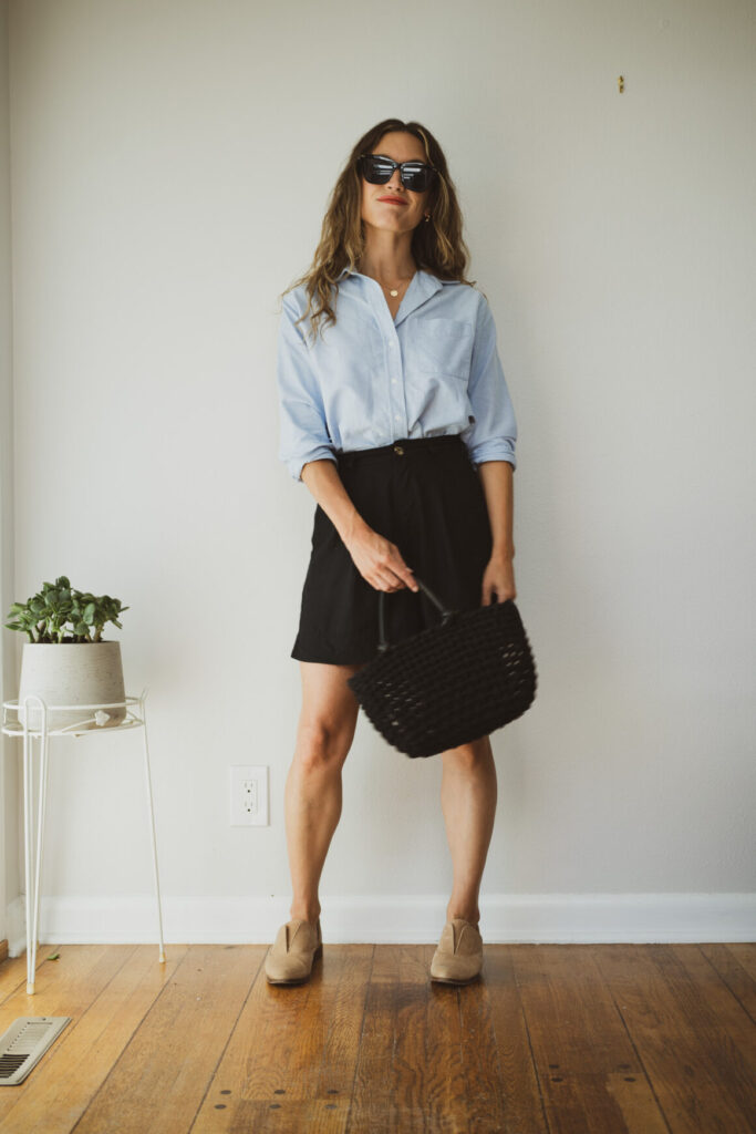 A person with wavy hair, wearing a light blue linen button-down shirt with rolled sleeves and black tailored shorts. They have a black woven tote bag and are wearing beige loafers. They are accessorized with tortoiseshell-style sunglasses and a gold necklace, embodying 'The Coastal Heiress' look from The Coastal Heiress.jpg.