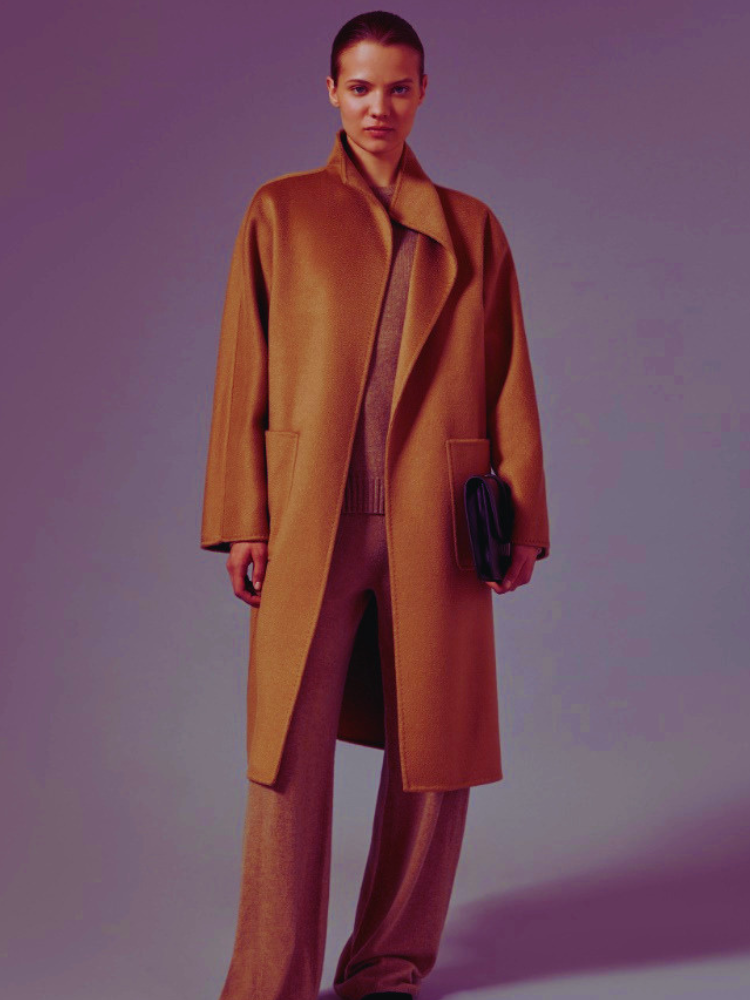 A woman posing in a studio against a neutral background, showcasing a monochromatic old money outfit from The Monochromatic Muse All-Camel Everything (2).jpg. She is wearing a long, structured camel wool coat over a matching knit sweater and fluid wide-leg knit trousers, while carrying a dark leather clutch purse.