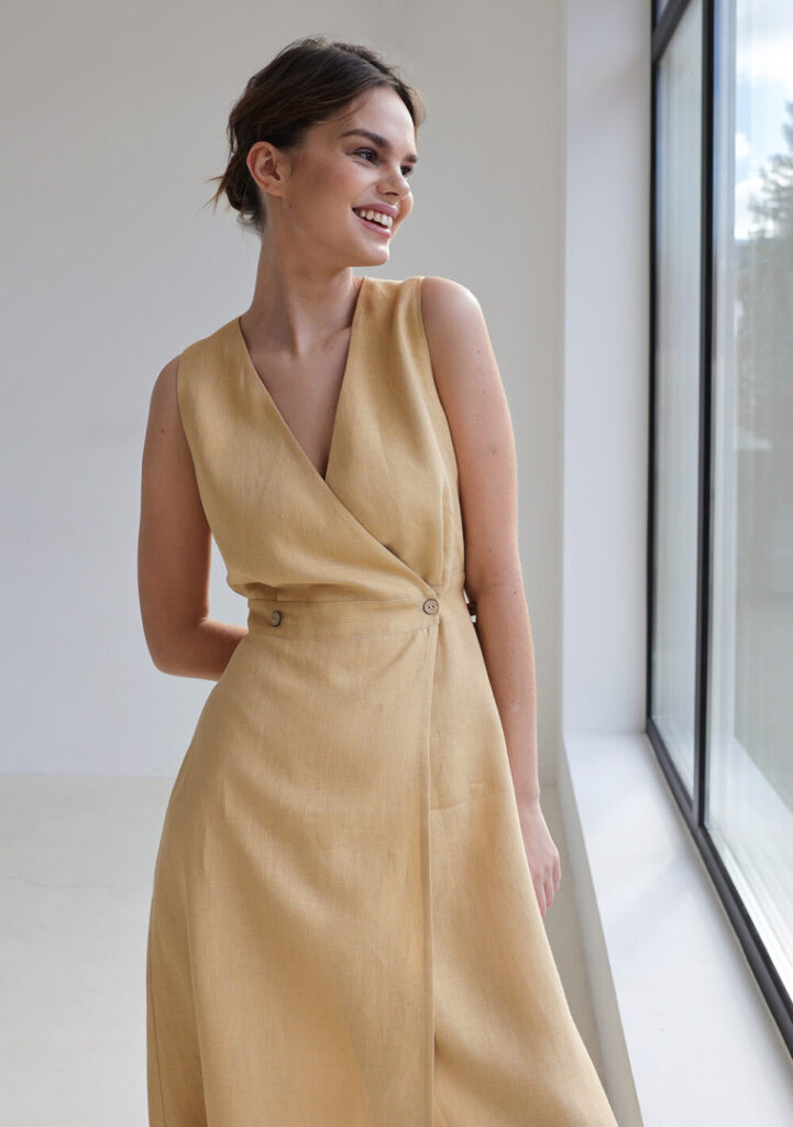 A smiling brunette woman with her hair tied back in an updo, wearing a chic minimalist sleeveless linen wrap dress in a light beige-yellow color with button details at the waist, modeling the old money Riviera look from linen-dress-riviera.jpg.