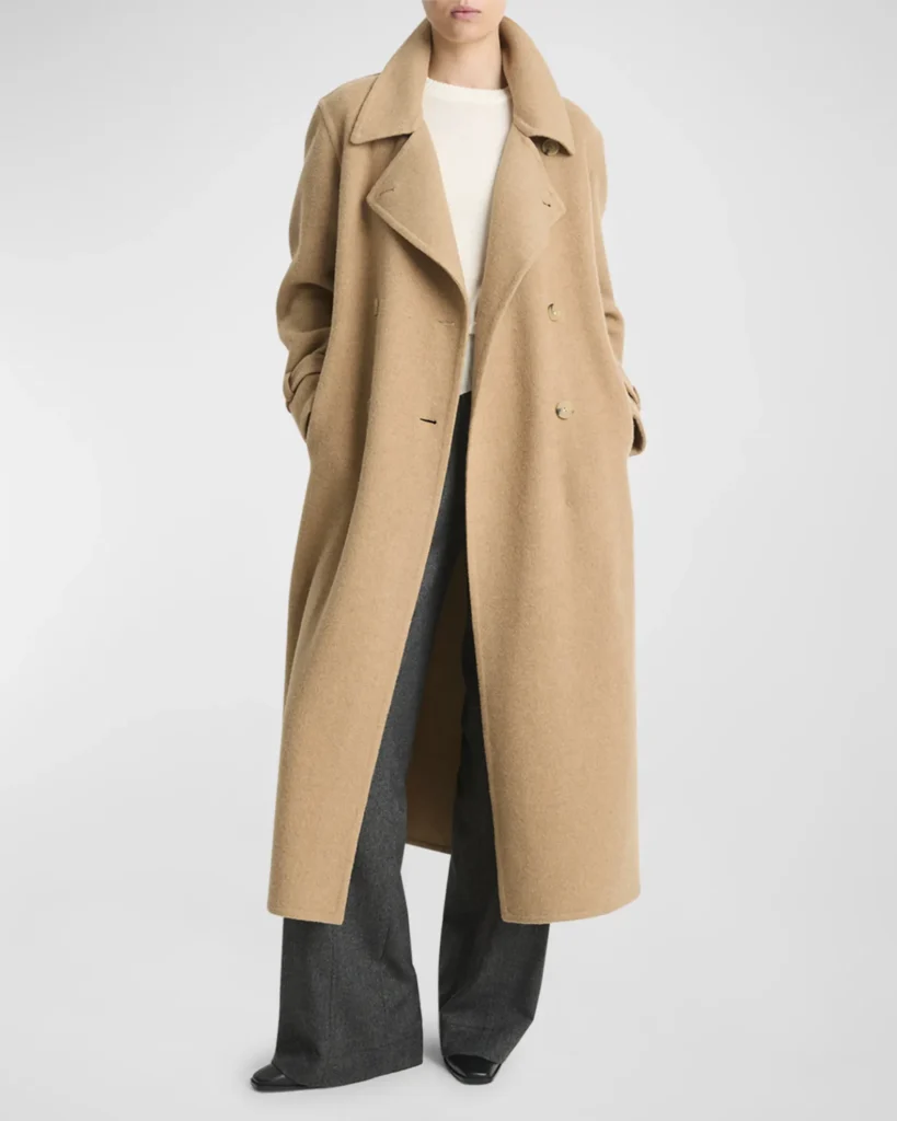 A full-length fashion photograph of a person from the chin down, standing against a plain, light-gray background. They are wearing a long, oversized camel-colored wool trench coat with large lapels and button details, layered over a cream-colored top and wide-leg, dark gray trousers. Their hands are tucked into the coat pockets, and the tips of black pointed-toe shoes are visible at the bottom.